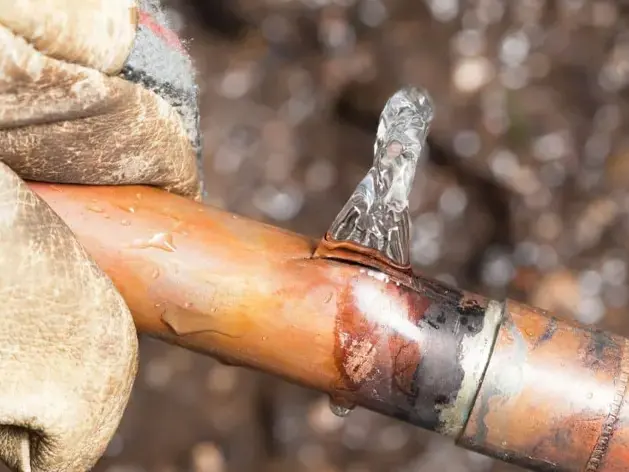 Gloved hand holding a split copper supply pipe with water spraying from the break